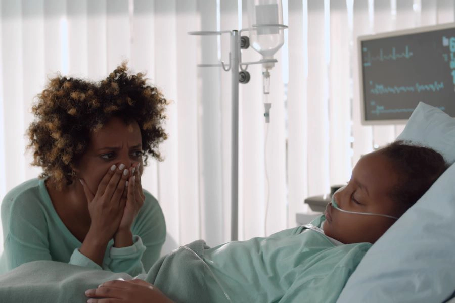 Upset young african woman crying sitting near little daughter sleeping in bed at hospital